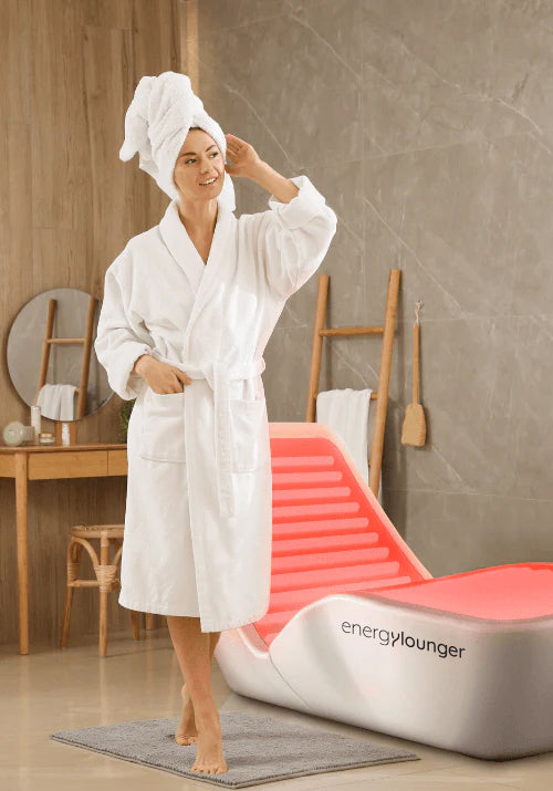 Person in a white robe standing beside the EnergyLounger with the blanket in a warm spa bathroom environment.