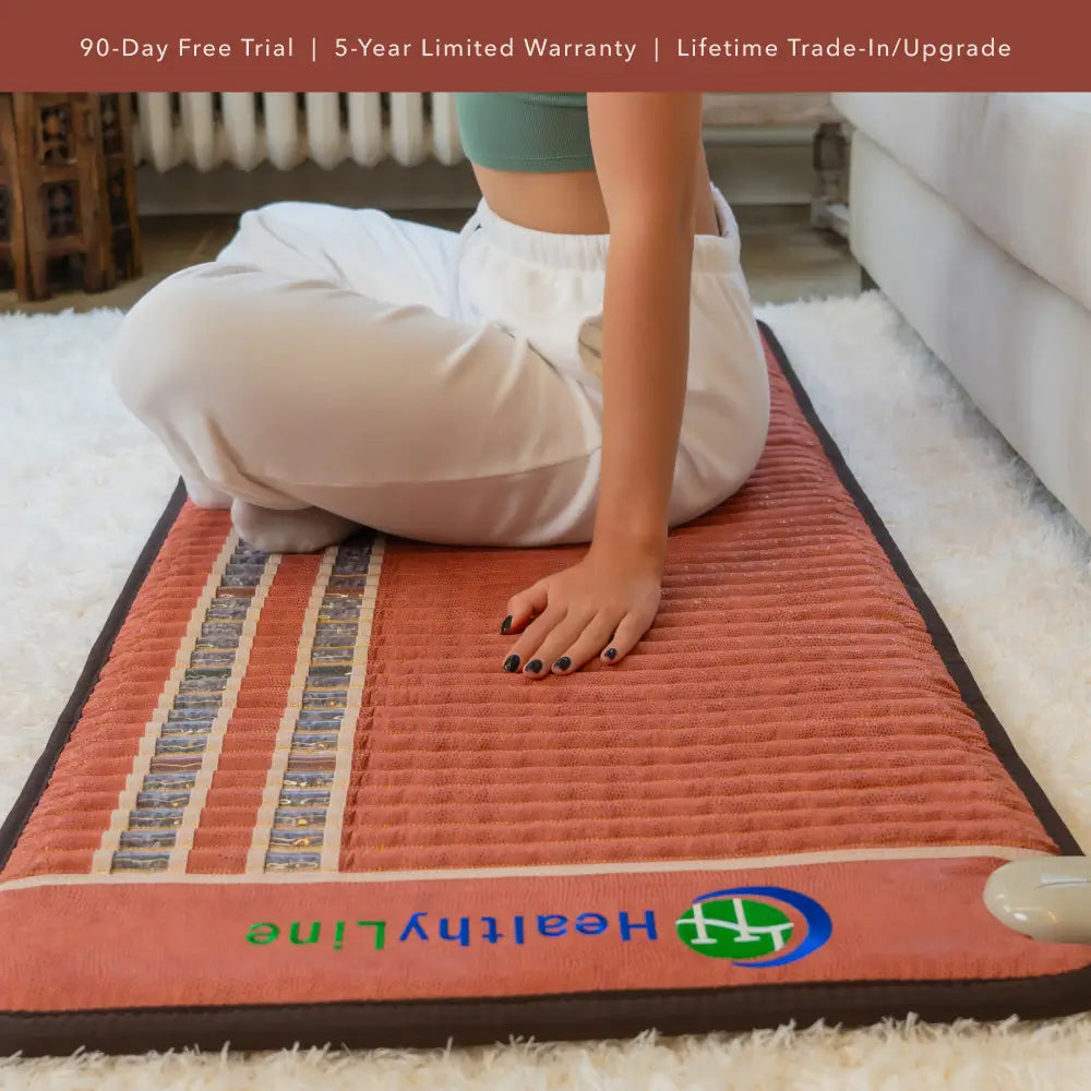 “Person sitting cross-legged on a full-length HealthyLine TAO heating mat in a living room setting, showing the textured gemstone surface and logo at the foot of the mat.”
